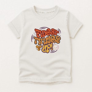 Puede incluir: Una camiseta de manga corta de color crema con un estampado gráfico en el pecho. El diseño incluye el texto "FIDGET THINGS UP!" en letras de burbuja naranjas y marrones, acompañado de un pequeño fidget spinner naranja y una cara sonriente amarilla.