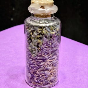 Może przedstawiać: Mała, przezroczysta szklana butelka wypełniona suszonymi pąkami lawendy i kilkoma białymi kryształkami. Butelka ma korek z jasną, teksturowaną górą. Tło to jednolita fioletowa powierzchnia.