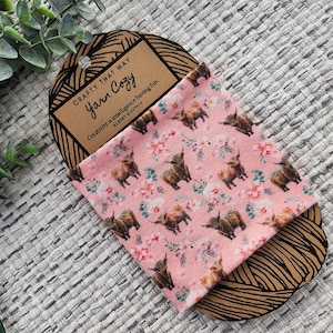 Op de afbeelding: Een roze stof met een patroon van bruine Highland-koeien en witte bloemen. De stof is gewikkeld om een ​​bruine papieren rol met de tekst "CRAFTY THAT WAY Yarn Cozy" en een citaat van Albert Einstein: "Creativiteit is intelligentie die plezier heeft."