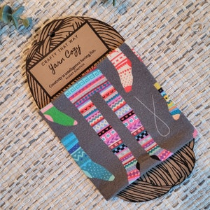 May include: A grey Yarn Cozy featuring colourful sock patterns. The socks display various designs, including geometric shapes and patterns in shades of pink, blue, green, and red. The tag reads "Crafty That Way" and "Yarn Cozy".