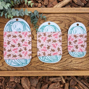 Könnte beinhalten: Drei rosa Stoff-Geschenkpapierquadrate mit einem Lebkuchenmann-Muster. Der Stoff ist rosa mit braunen Lebkuchenmännern und dem Text "Oh Snap!" und "Ideus 40" in rot gedruckt.