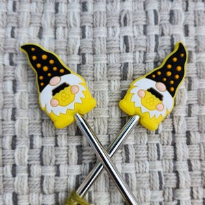 Puede incluir: Dos adornos para agujas de tejer con forma de gnomo. Los gnomos tienen sombreros negros con lunares naranjas, barbas blancas y cuerpos amarillos. Las agujas de tejer son plateadas y están cruzadas. El fondo es una tela gris claro texturizada.