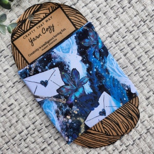 May include: A blue and white fabric cup cosy with a floral design and a quote from Albert Einstein: "Creativity is intelligence having fun." The cosy is packaged with a brown paper tag that says "Crafty That Way Yarn Cozy".