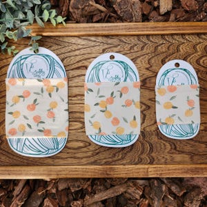 May include: Three white fabric yarn bowl cozies with a peach and leaf pattern. The cozies are in different sizes and are displayed on a wooden tray. The cozies are labelled "Crafty That Cozy" with a website address.