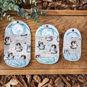 May include: Three different sizes of fabric cup cozies with a penguin and snowflake design. The cozies are beige with brown penguins and blue snowflakes. The text "Crafty Gray" is on the white tag.