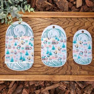 May include: Three fabric items with a winter pattern of penguins, Christmas trees, and gifts. Each is attached to a white card with a teal design and the text "Crafty Way". The items are displayed on a wooden tray.
