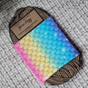 May include: A brown paper card with the text "CRAFTY THAT WAY Yarn Cozy" and the quote "Creativity is intelligence having fun. - Albert Einstein". The card is wrapped in a fabric with a rainbow mermaid scale pattern.