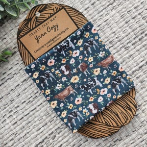 以下が含まれることがあります： 白と黄色の花柄と、茶色と黒の牛が繰り返し描かれた青い布。布は折り畳まれ、茶色の紙タグに「Crafty That Way Yarn Cozy Creativity is intelligence having fun. Albert Einstein」というテキストが書かれています。