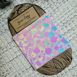 May include: A gray fabric with a colorful abstract pattern of pink, blue, and yellow circles. The fabric is folded and placed on top of a brown paper label with the text "CRAFTY THAT WAY Yarn Cozy" and a quote by Albert Einstein: "Creativity is intelligence having fun."