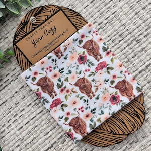 以下が含まれることがあります： 花柄と茶色のハイランド牛のイラストが描かれた白い生地。生地は、"Crafty That Way Yarn Cozy"というテキストと、アルバート・アインシュタインの創造性に関する引用が書かれた茶色の紙のカードに巻かれています。