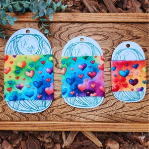 May include: Three colorful fabric items with a watercolor heart pattern in shades of red, blue, yellow, and green. Each item is attached to a white card with a logo and website address. The items are displayed on a wooden surface.