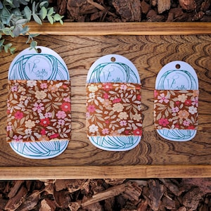 May include: Three oval-shaped fabric gift tags with a floral pattern in shades of brown, pink, and cream. The tags are labelled "Crafty" and feature a stylised needle and thread design.