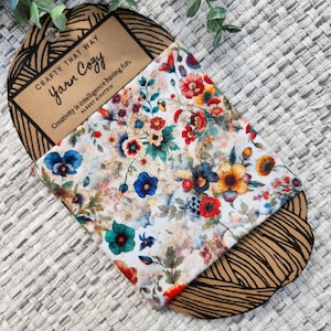 May include: A floral patterned Yarn Cozy, featuring a variety of colorful flowers in shades of red, blue, yellow, and teal on a white background. The product is packaged with a brown tag that reads "Crafty That Way Yarn Cozy" and a quote by Albert Einstein.