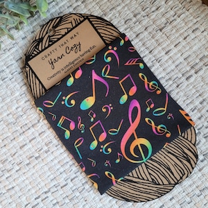 May include: A black fabric with a rainbow-colored musical note pattern. The fabric is folded and placed on top of a brown paperboard with the text "CRAFTY THAT WAY Yarn Cozy Creativity is intelligence having fun. ALBERT EINSTEIN" printed on it.