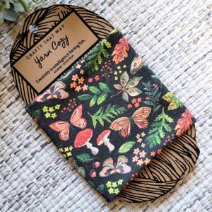 May include: A black fabric pouch with a colourful pattern of butterflies, leaves, and mushrooms. The pouch is folded and sitting on a brown cardboard tag with the text "Crafty That Way Yarn Cozy" and a quote by Albert Einstein: "Creativity is intelligence having fun."