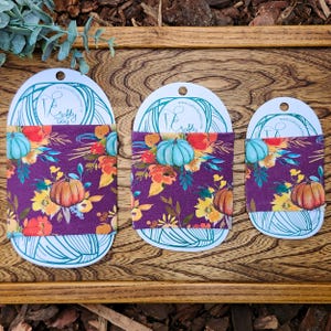 May include: Three fabric drink sleeves with a fall design. The sleeves have a dark purple background with orange, yellow, and teal pumpkins and floral patterns. Each sleeve is attached to a white card with the text "crafty the way".