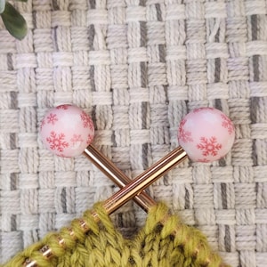 May include: Close-up of knitting needles with pink snowflake-decorated tops. The needles are crossed over a section of green knitted fabric. The needles are copper-colored, and the background is a woven, light gray fabric.