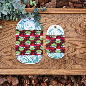 May include: Two Grinch-themed fabric cup cozies, one larger and one smaller, on a wooden surface. The cozies feature a red and black plaid pattern with a repeating image of the Grinch wearing a Santa hat.