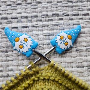 May include: Two knitting needles with blue gnome-shaped stitch markers. The needles are crossed and are holding a green knitted fabric.