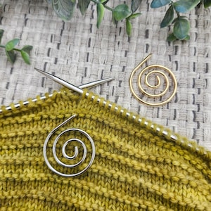 May include: Two silver and gold spiral stitch markers on a yellow knitted fabric. The stitch markers are used to mark the beginning or end of a row of knitting.