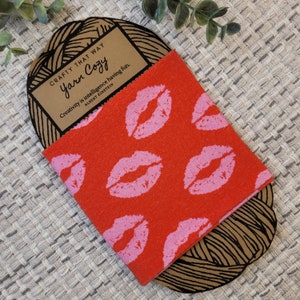 May include: A red fabric swatch with a pink lip print pattern. The fabric is wrapped around a brown yarn ball with the text "CRAFTY THAT WAY Yarn Cozy Creativity is intelligence having fun. ALBERT EINSTEIN" printed on a label.
