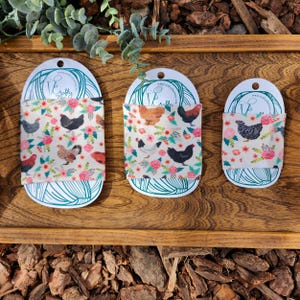 May include: Three different sizes of fabric coffee cup sleeves with a floral pattern and chickens. The sleeves are white with pink flowers and brown, black, and white chickens. The text "Crafty Threads" is on each sleeve.