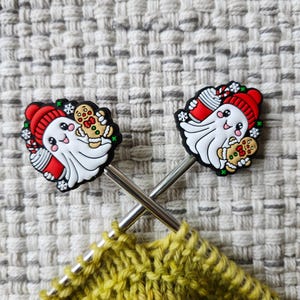 May include: Two Christmas-themed stitch markers featuring a smiling ghost wearing a red hat and holding a gingerbread man. The markers are on silver metal needles, with a yellow knitted piece in the foreground. The markers are black, white, red, and green.