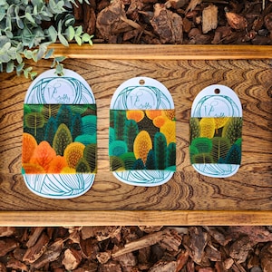 Op de afbeelding: Drie verschillende maten herbruikbare voedselverpakkingen met een bosprintontwerp. De verpakkingen zijn gemaakt van bijenwas, biologisch katoen en boomhars. De verpakkingen worden getoond op een houten oppervlak.