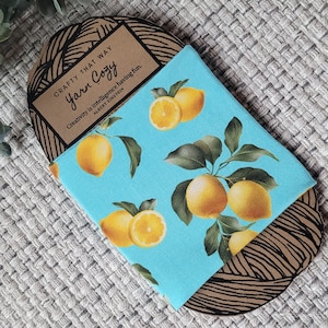 May include: A blue fabric yarn cosy with a lemon pattern, featuring whole and sliced lemons with green leaves. The cosy is partially encased in a brown cardboard holder with the text "Crafty That Way Yarn Cozy" and a quote by Albert Einstein.