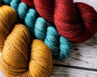 May include: Three skeins of yarn in different colours: a deep red, a teal blue, and a golden yellow. The yarn is arranged in a loose, overlapping pattern.