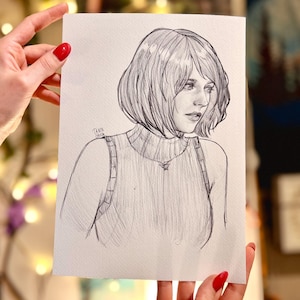 May include: A pencil sketch portrait of a person with short hair, wearing a sweater. The artwork is held by hands with red nail polish. The artist's signature is visible in the lower left corner.