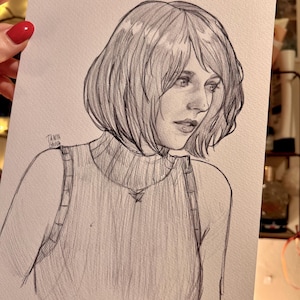May include: A pencil sketch portrait of a woman with short hair, wearing a turtleneck sweater. The artwork is signed "Tanya Gana" in the lower left corner. The drawing is on white paper.