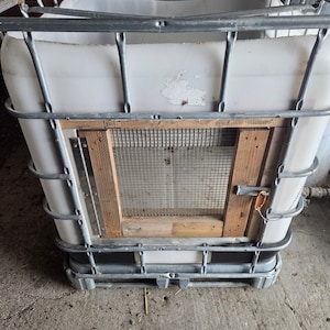 May include: A large, white plastic container with a metal cage frame. A wooden door with wire mesh is attached to the front. The container sits on a metal pallet base, suggesting industrial or agricultural use.