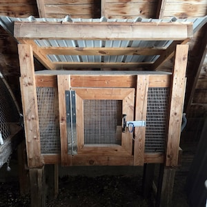 Puede incluir: Un conejar de madera con techo de metal, laterales de malla metálica y puerta con bisagras. El conejar está elevado sobre postes de madera y tiene un pestillo para asegurar la puerta. La estructura es de madera natural.