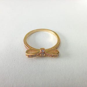 May include: A gold ring with a bow-shaped design. The bow and the ring band are embellished with small, clear gemstones. The ring is set against a white backdrop, accentuating its intricate details and elegant design.
