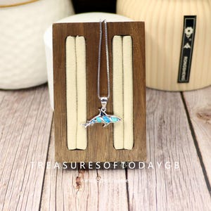 Può includere: Una collana con ciondolo a forma di squalo argentato con dettagli in opale blu. Il ciondolo a forma di squalo è esposto su un espositore di gioielli in legno. La collana ha una catena d'argento. Lo sfondo include una superficie in legno e un oggetto decorativo.
