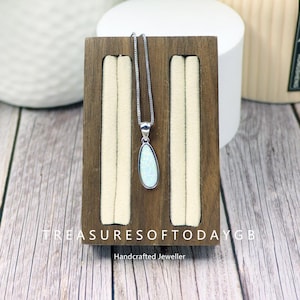 May include: A silver necklace featuring a pear-shaped, opalescent pendant. The pendant is presented on a wooden jewellery stand with cream inserts. The stand displays the text "TREASURESOFTODAYGB" and "Handcrafted Jeweller".