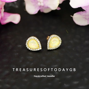 May include: Gold-toned teardrop earrings with a central opal-like stone, surrounded by a halo of small, clear crystals. The earrings are set against a dark background, with the text "TREASURESOFTODAYGB" and "Handcrafted Jeweller" visible.