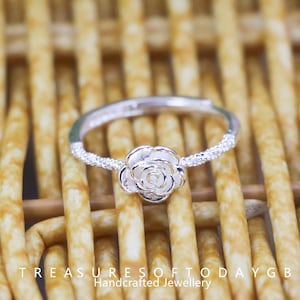 Puede incluir: Anillo ajustable plateado con un centro en forma de rosa. La banda del anillo está adornada con pequeños detalles brillantes. El anillo se muestra sobre una superficie tejida de color marrón claro. El texto "TREASURESOFTODAYGB Handcrafted Jewellery" está en la parte inferior.