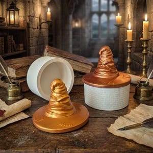 May include: A gold-colored Sorting Hat-shaped container with a white brick-like base and lid. The lid is off, revealing the hat's interior. The container sits on a wooden surface with parchment and candles.