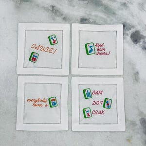 May include: Four white square coasters with embroidered Mahjong tiles and phrases in orange and red thread. The phrases include "PAUSE!", "bird bam cheers!", "everybody loves a", and "BAM DOT CRAK".