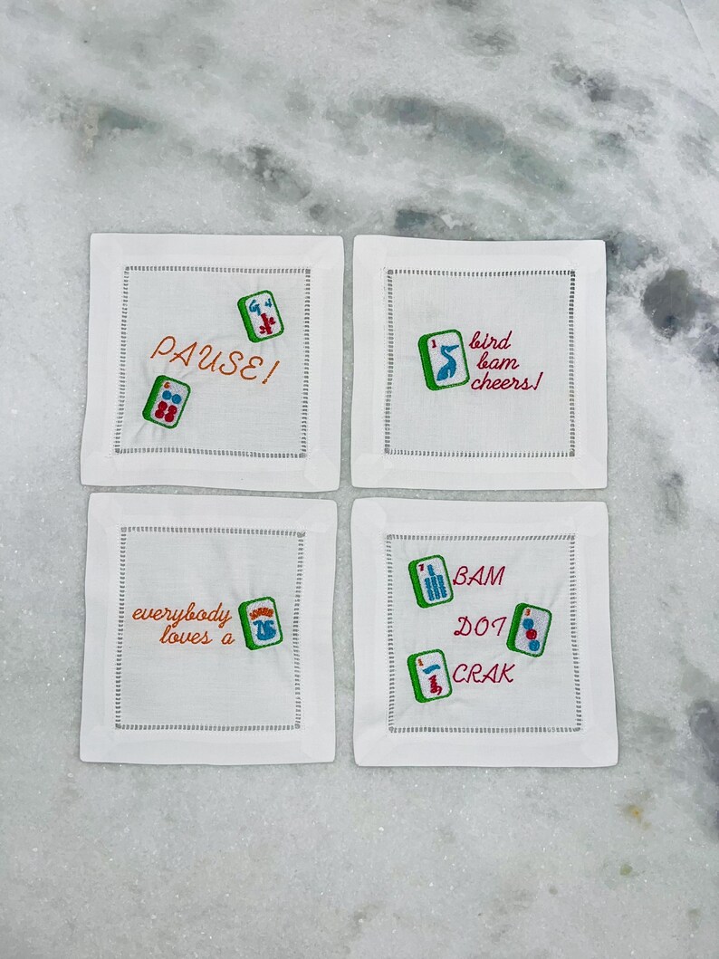 May include: Four white square coasters with embroidered Mahjong tiles and phrases in orange and red thread. The phrases include "PAUSE!", "bird bam cheers!", "everybody loves a", and "BAM DOT CRAK".