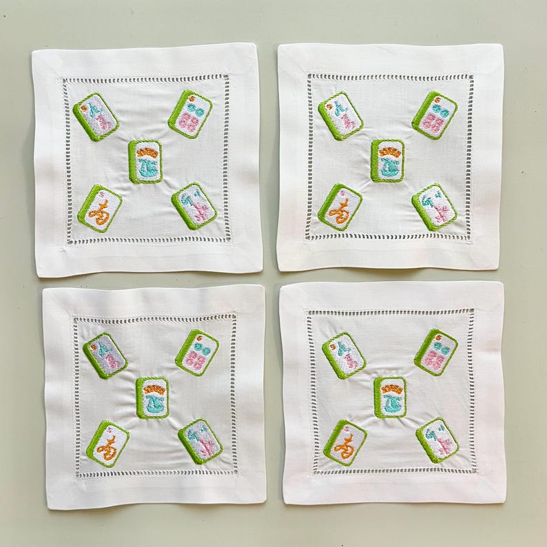 May include: Set of four square white linen coasters with embroidered Mahjong tile designs. Each coaster features a green border and colorful tile images, perfect for protecting surfaces from drink condensation.