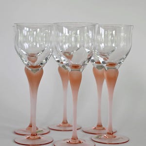 Puede incluir: Un juego de cinco copas de vino de cristal transparente con tallos altos y delgados. Cada tallo presenta una base rosada esmerilada y acanalada que se funde con un cáliz liso y transparente. Las copas se apoyan sobre bases circulares rosadas.