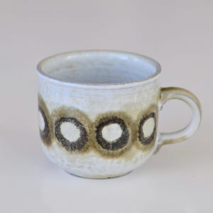 Puede incluir: Taza de cerámica color crema con una serie de patrones circulares marrones y negros. La taza tiene una forma redondeada con un asa, adecuada para bebidas calientes. El diseño es simple y rústico.