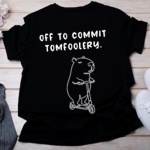 Może przedstawiać: Czarny t-shirt z napisem "OFF TO COMMIT TOMFOOLERY." Poniżej tekstu znajduje się biały rysunek konturowy kapibary na skuterze. Koszulka wykonana jest z miękkiego materiału.