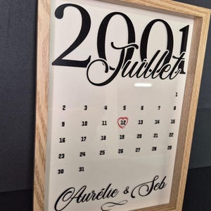 Peut inclure: Impression encadrée d'un calendrier de juillet 2001, avec le 12e jour mis en évidence par un cœur rouge. Les noms "Aurélie & Seb" sont écrits en bas. Le cadre est en bois clair. Le texte est en script noir.