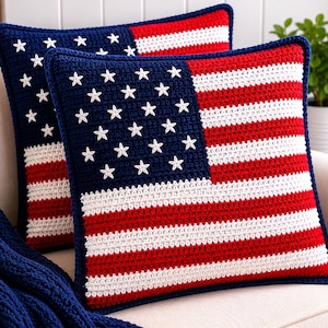May include: Two square crochet pillows with a design resembling the American flag. The pillows feature a blue field with white stars and red and white stripes. The pillows are on a cream-colored surface.