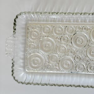 Może przedstawiać: Prostokątna, przezroczysta szklana taca do serwowania z wzorem koncentrycznych okręgów. Taca ma falisty brzeg i małe uchwyty. Projekt przedstawia mieszankę dużych i małych okręgów.