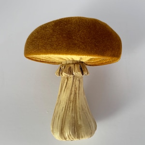 Pode incluir: Um cogumelo decorativo com um chapéu castanho-dourado texturizado e um caule creme. O chapéu tem uma aparência aveludada e o caule tem um design esculpido detalhado. Esta peça lúdica adiciona um toque de decoração inspirada na natureza.